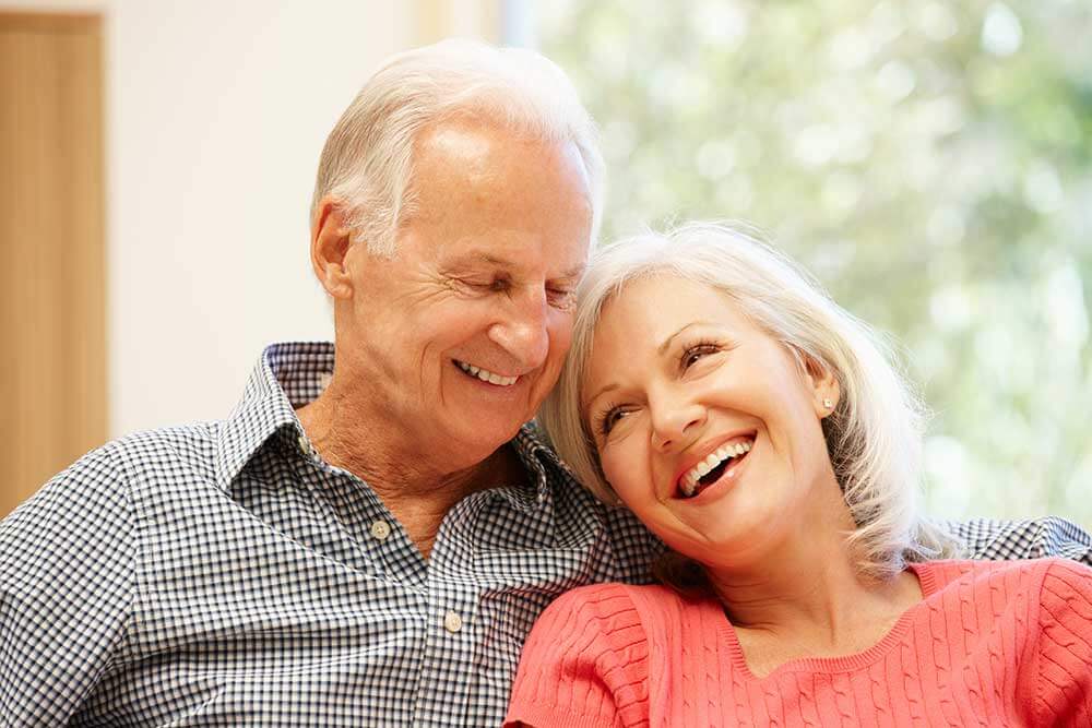 An older couple is smiling and leaning on each other. Their restorative dentistry looks beautiful.