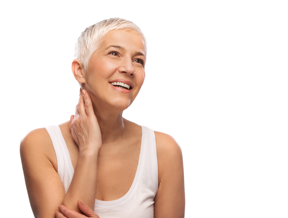 A woman places a hand on her neck and smiles. She's wearing white and has white hair and white restorative dentistry.
