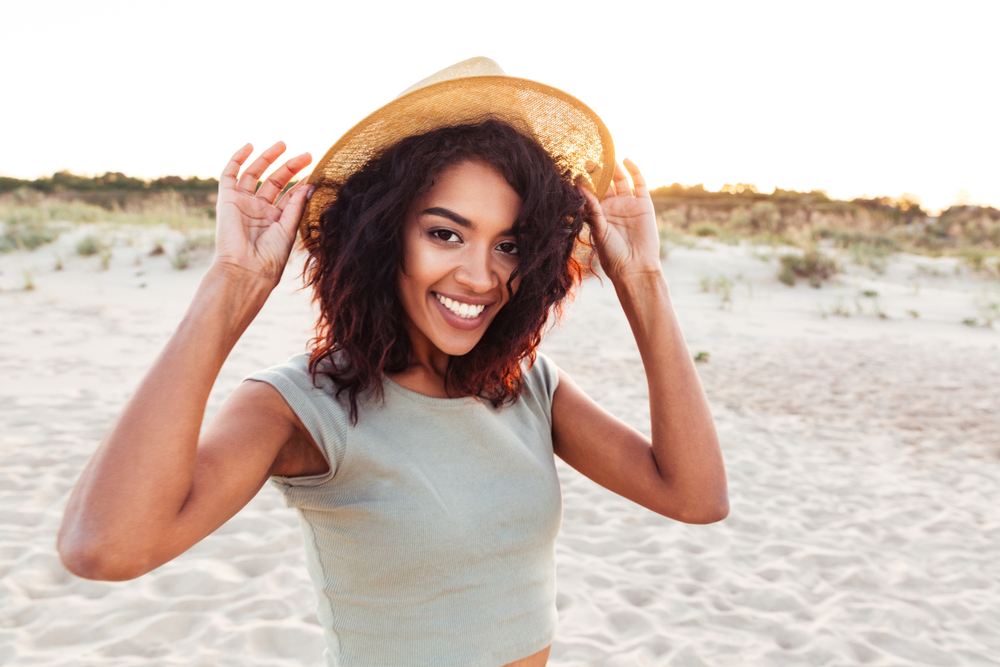 A woman on the beach wearing a hat is smiling comfortably thanks to periodontics.